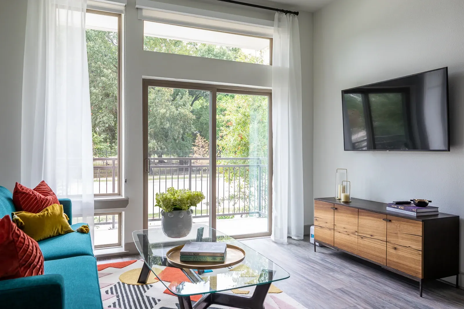 Modern living room with turquoise sofa, colorful rug, glass coffee table, and wall-mounted TV.