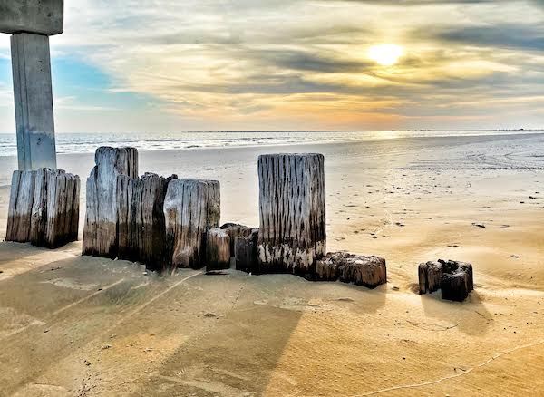 A pile of wooden posts on a beach with the sun shining through the clouds.