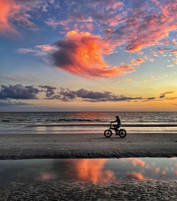 A person is riding a bike on the beach at sunset.