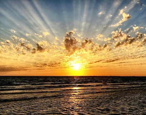 The sun is setting over the ocean and shining through the clouds.