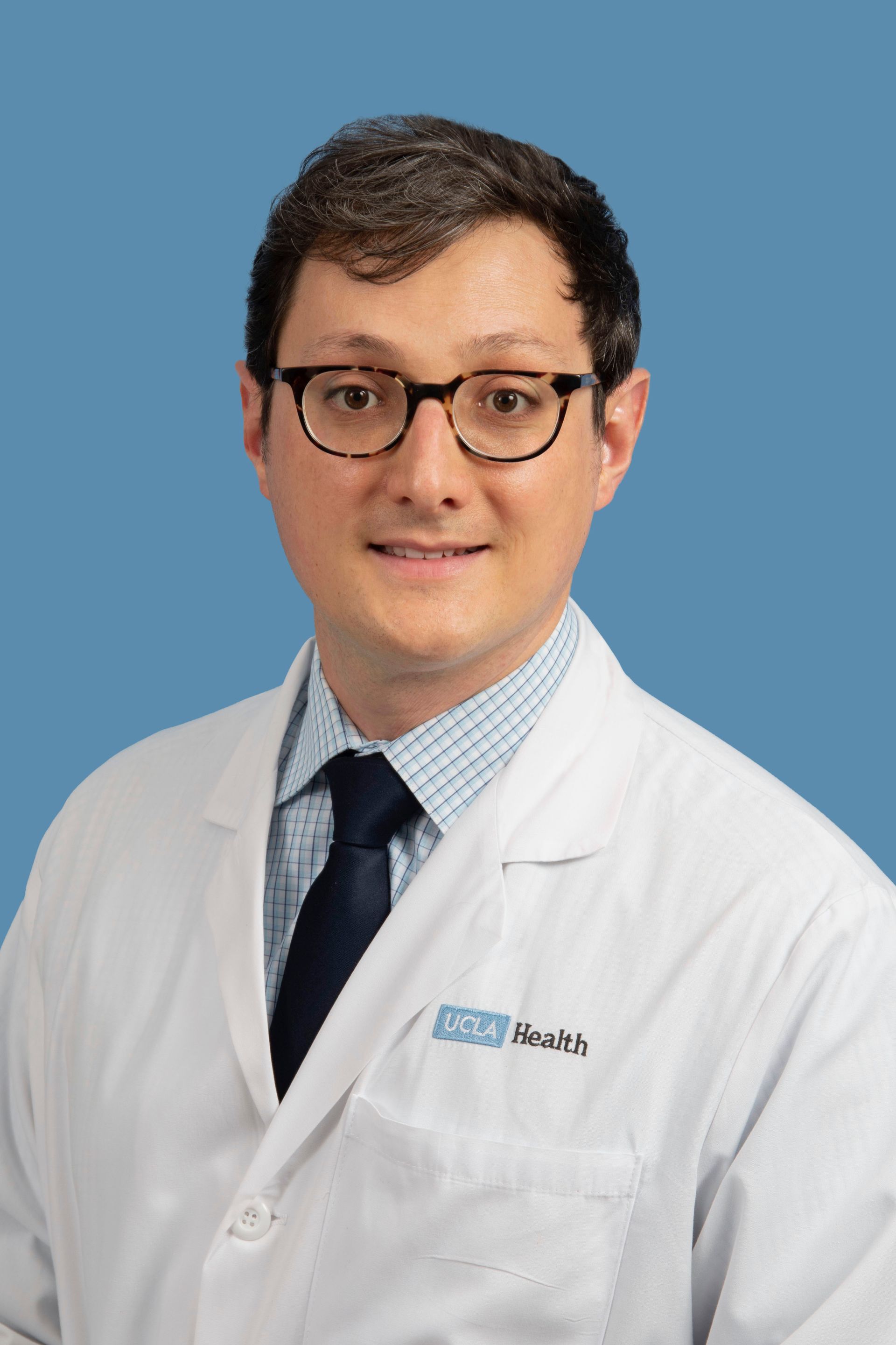 A man in a white lab coat and tie is standing in front of a blue background.