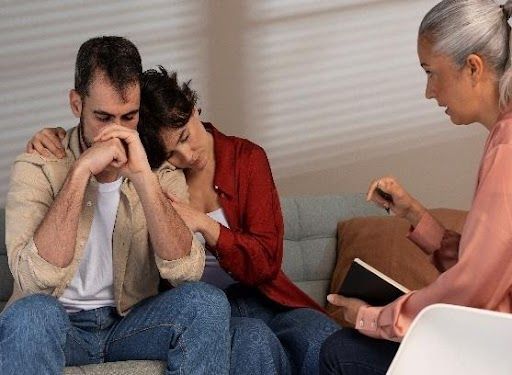 Couple in distress on a couch, comforted by each other and a therapist taking notes.