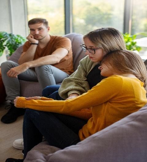 Two women embrace, comforted. A man watches from a separate chair. Interior, daytime.