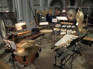 Percussion instruments, including cymbals, gongs, drums, and xylophone, arranged on stands in a concert hall.