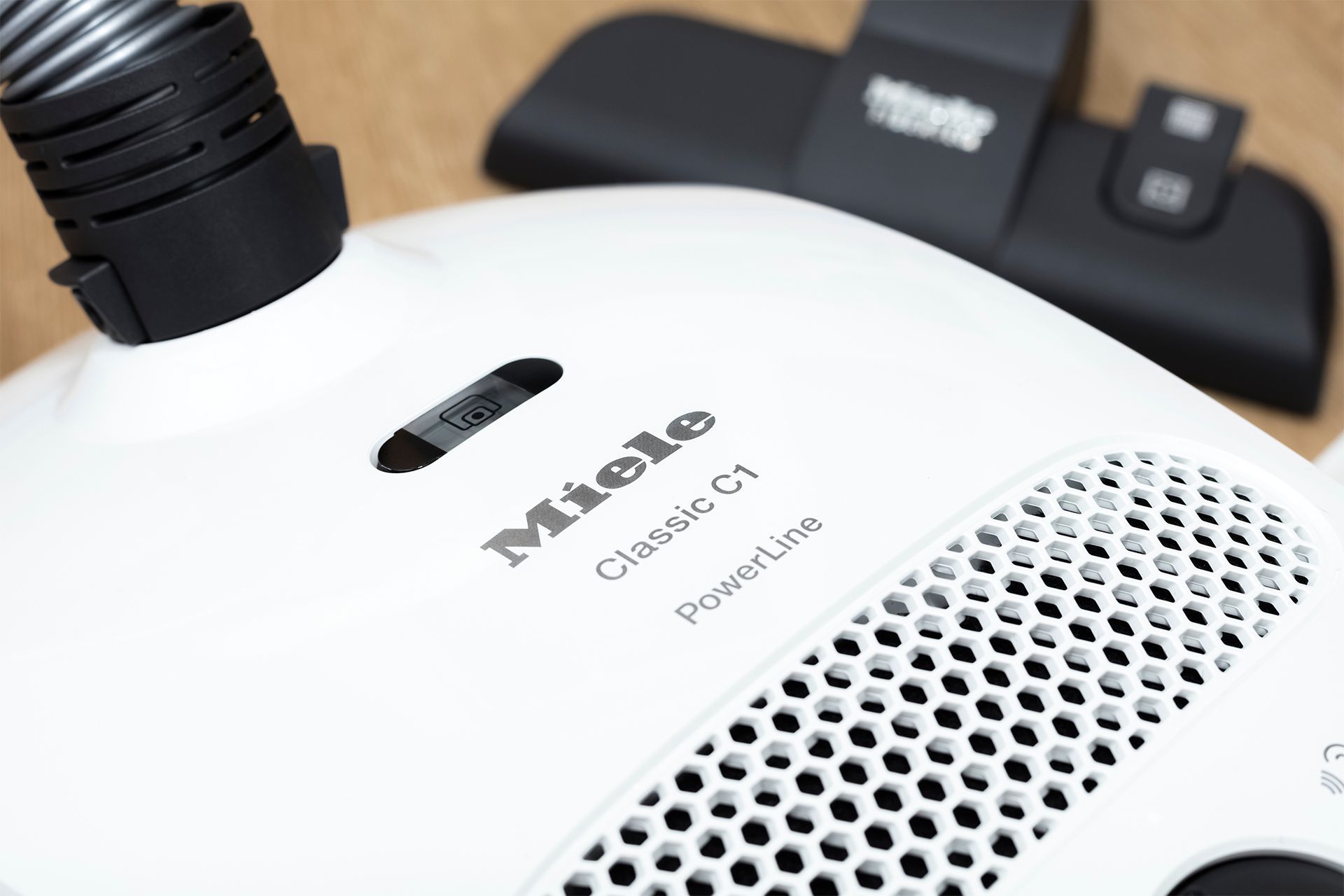 White Miele Classic C1 vacuum cleaner with hose and brush attachment, on a wood surface.