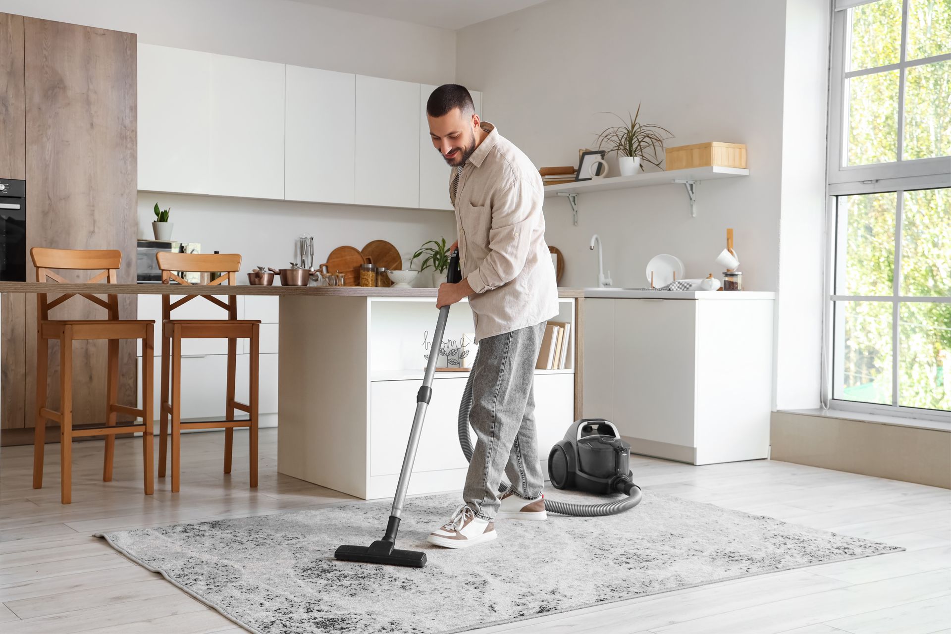 Man vacuuming a gray rug in a kitchen.