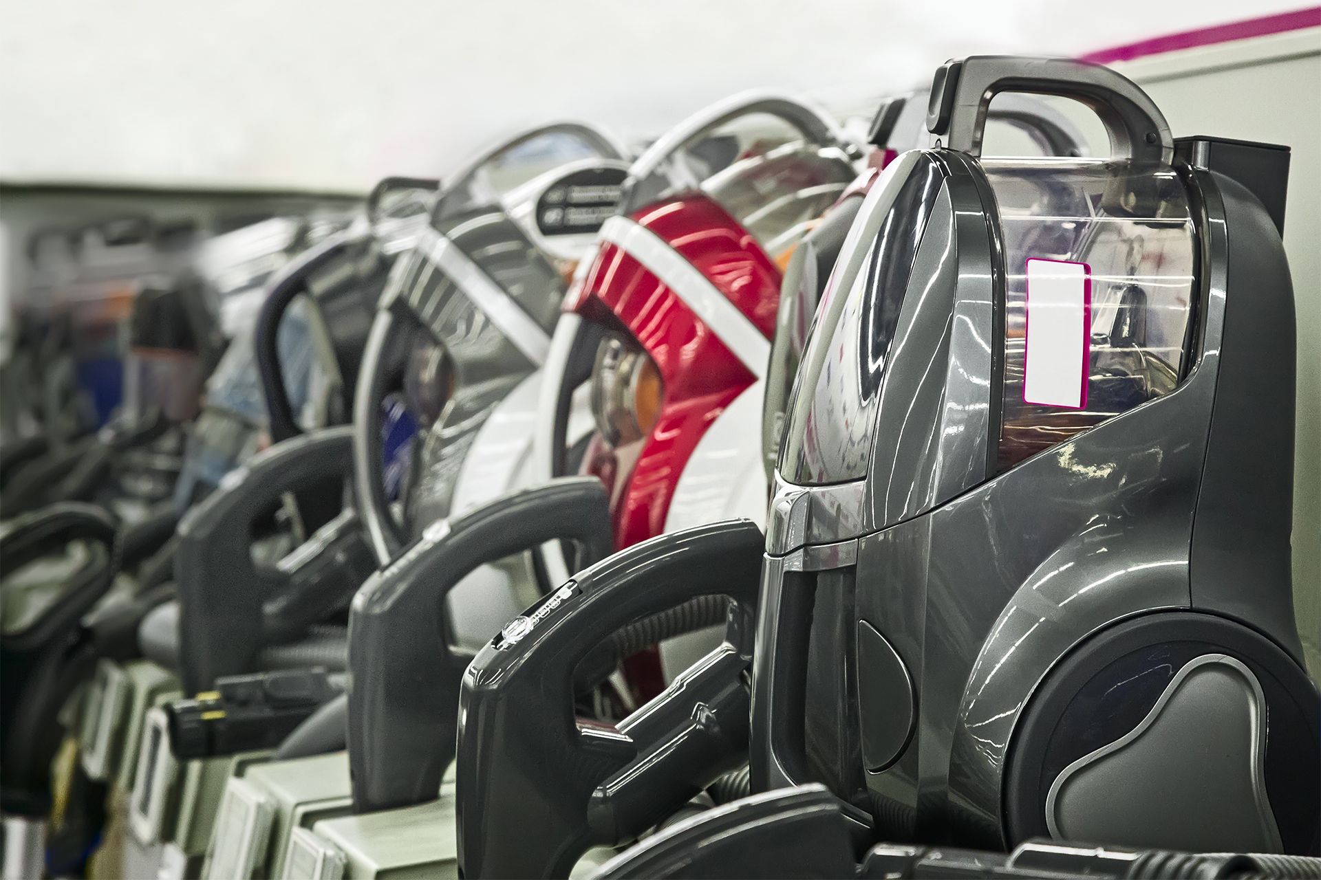 Row of black and gray vacuum cleaners on display in a store with a person in red and white attire in the background.
