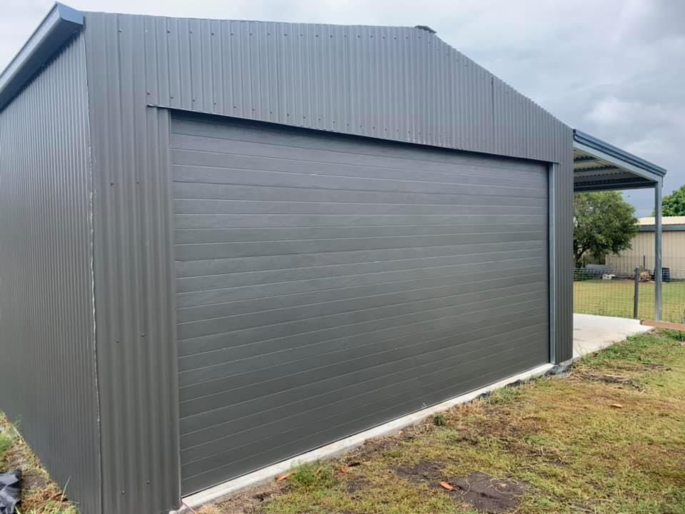 A gray garage with a sliding garage door is sitting in the middle of a grassy field.
