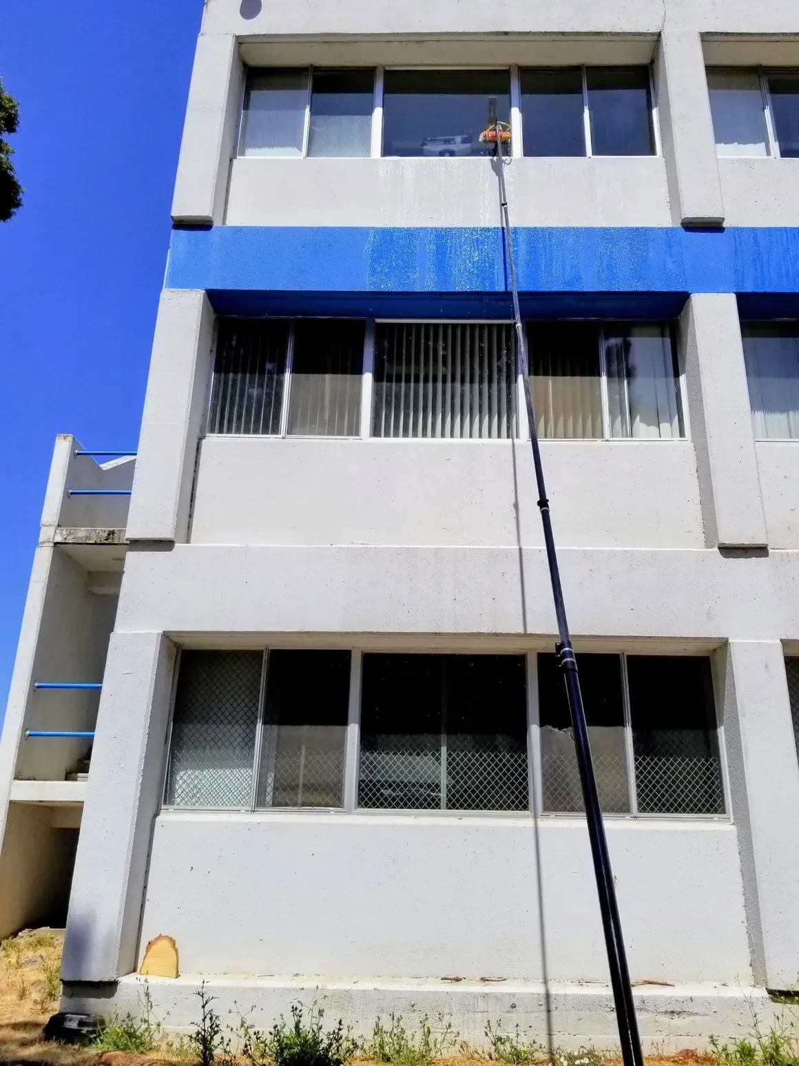A window cleaner using a long pole to wash windows on a tall, white building with blue accents.