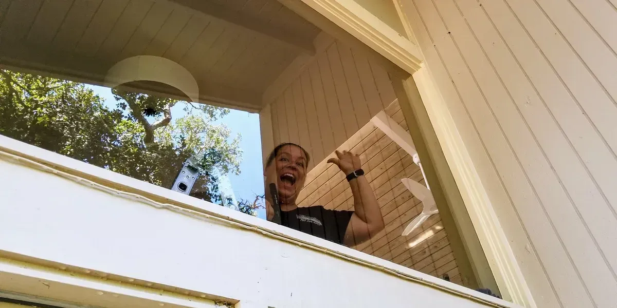 Woman smiling, giving a thumbs up from a window. Sunlight streams through.