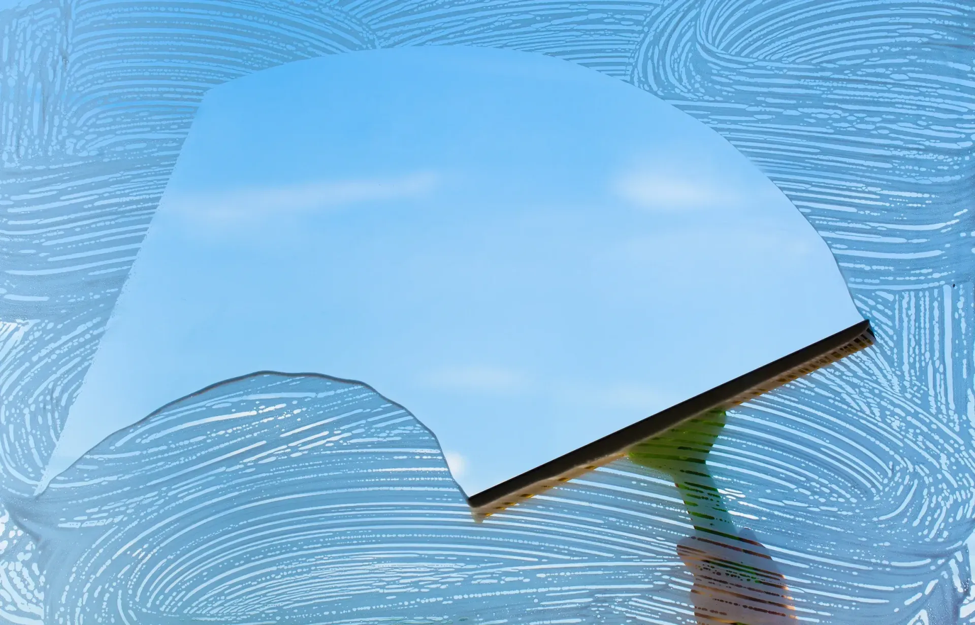 Hand using a squeegee to clean a window, revealing blue sky and clouds.