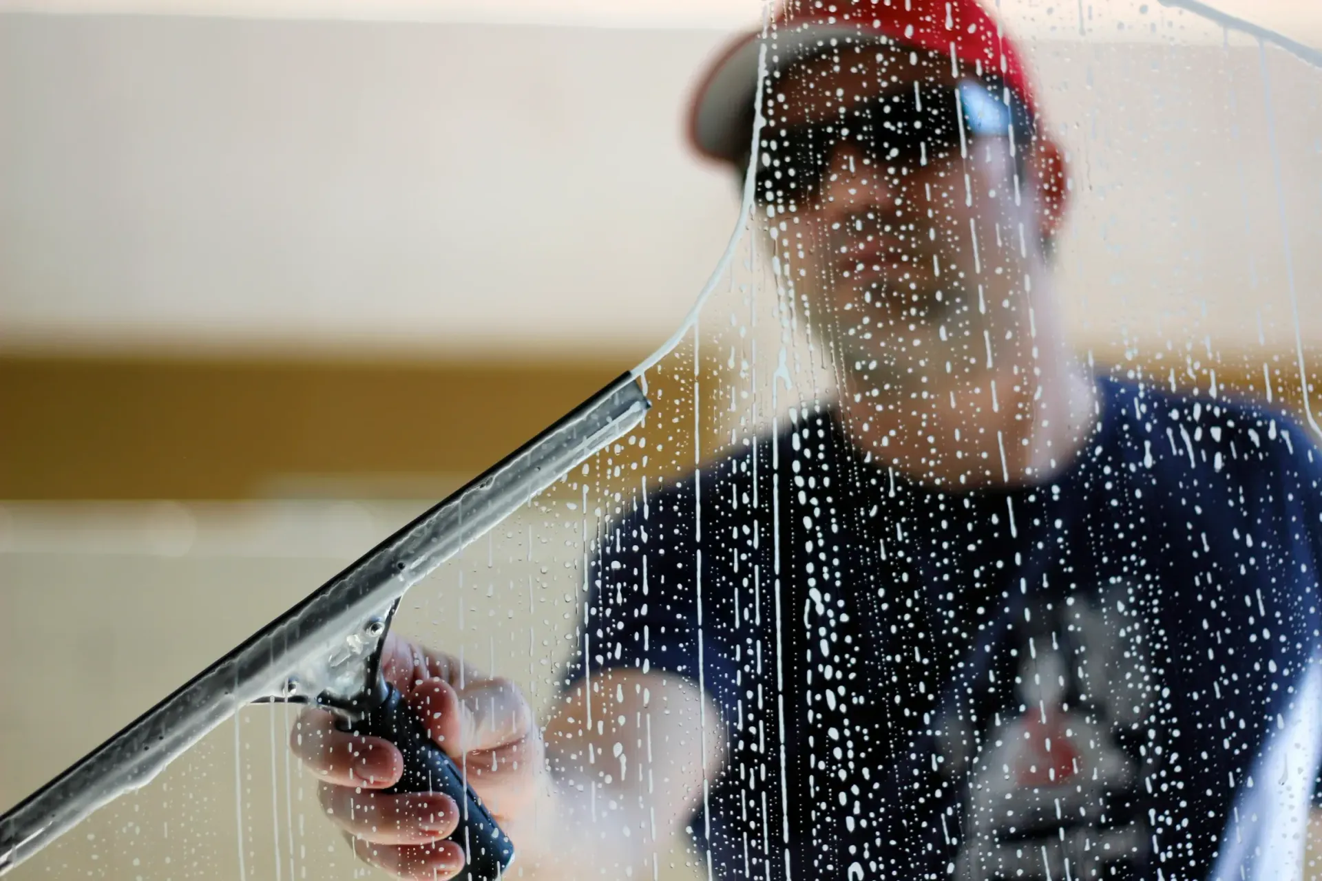 Man using a squeegee to clean a window, wearing a red cap and sunglasses.