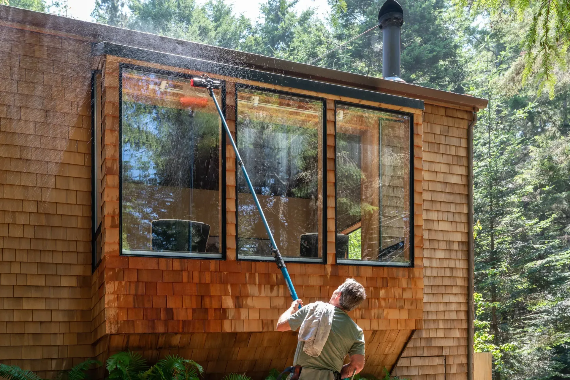 Person washing windows on a wood-shingled house with a long-handled brush on a sunny day.