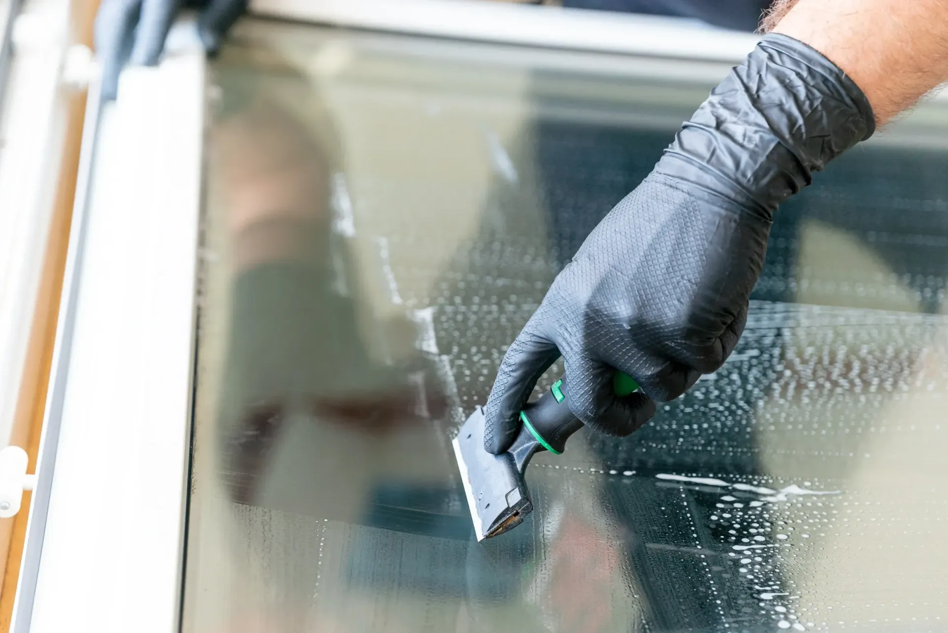 Person wearing black gloves scraping a window pane, applying cleaning solution.