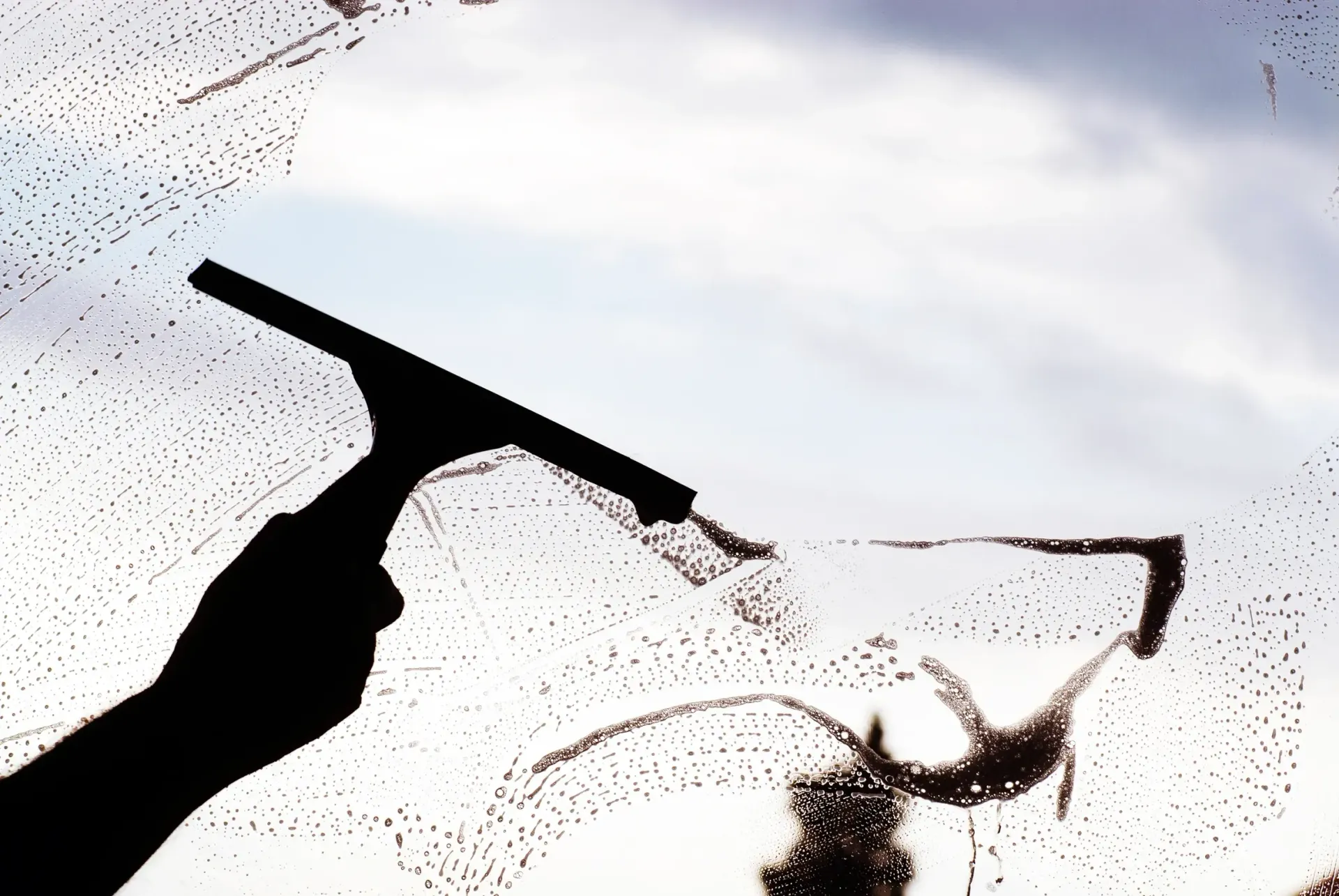 Silhouette of a hand using a squeegee to clean a window, sky visible through the clear portions.