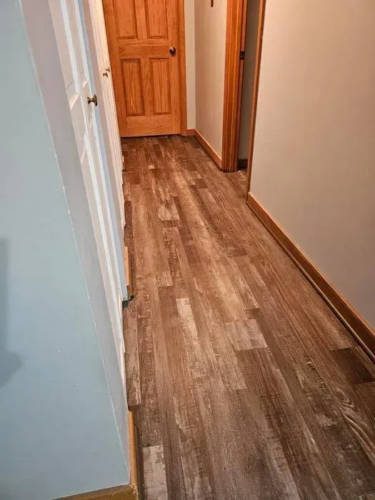 Hallway with wood-look flooring and doors. White cabinet on the left, brown door at the end. Gray walls.