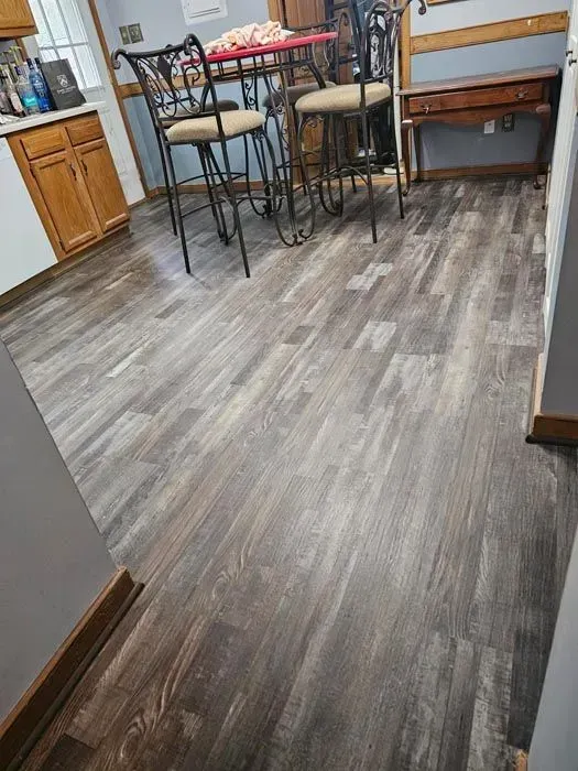 Wooden-look flooring in a kitchen area. Dark gray planks extend across the space, with a table and chairs in the background.