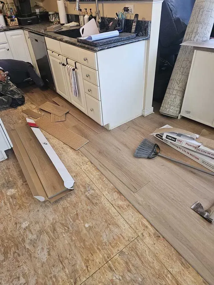 Kitchen renovation in progress: new flooring being installed over old, damaged subfloor. Cabinets and appliances are present.