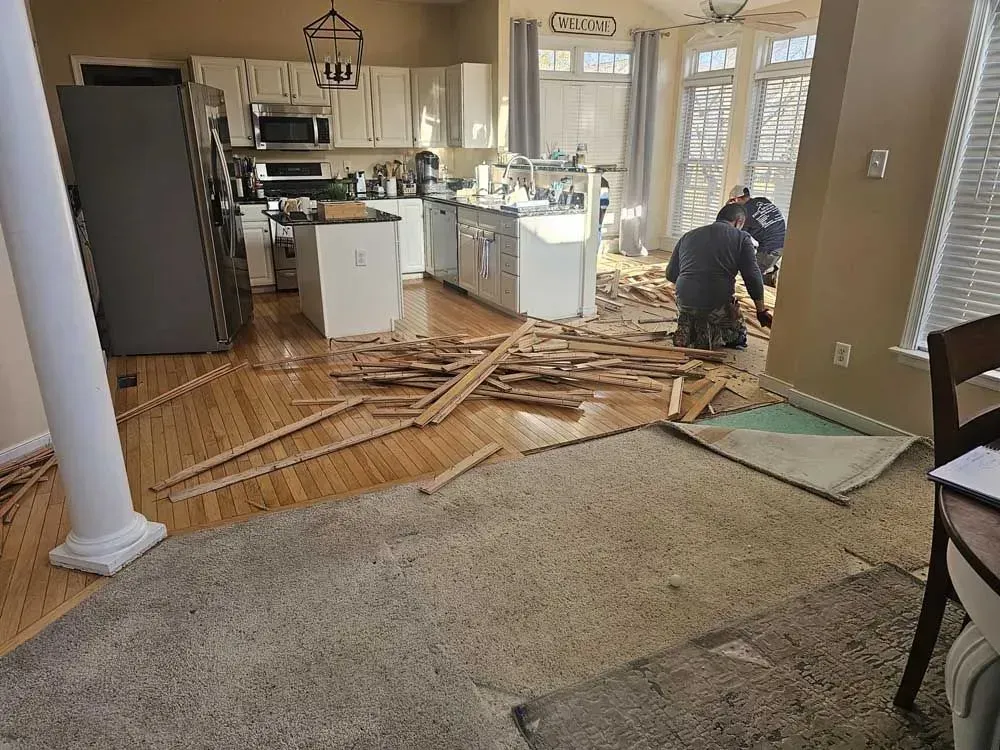Kitchen renovation: Workers removing hardwood flooring, debris scattered. Cabinets, fridge, and windows visible.