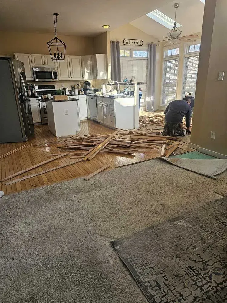 Hardwood floor being removed from a kitchen. Contractor working, debris scattered on floor and rug. Natural light.