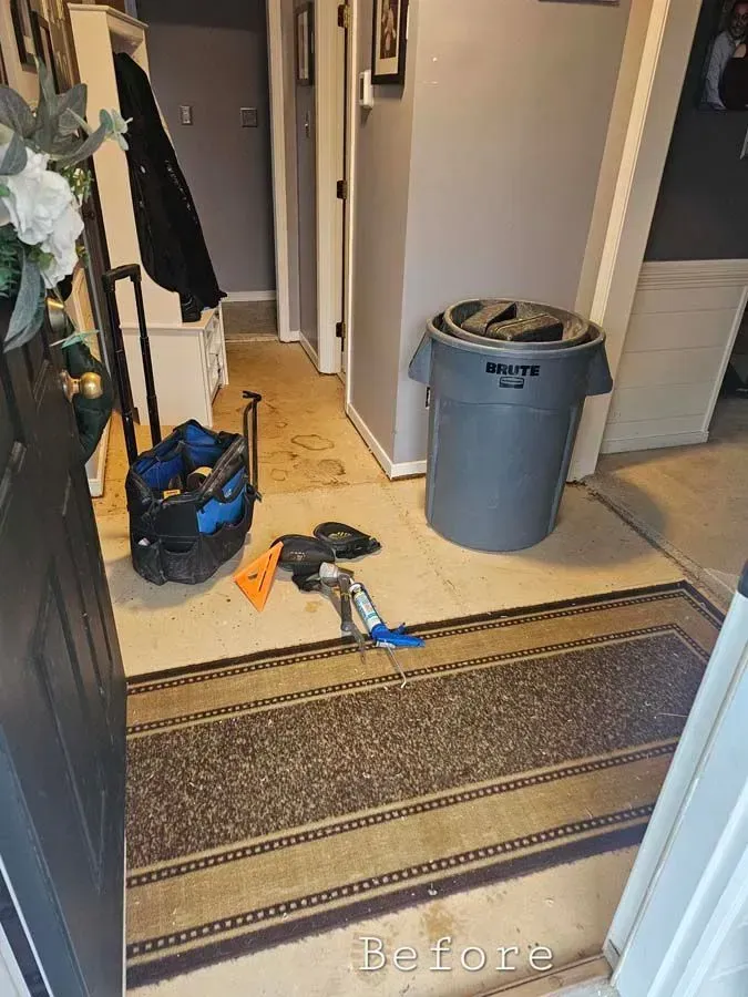 Entryway with work tools, trash can, and dirty rug. Dark door, light walls, and hallway visible.