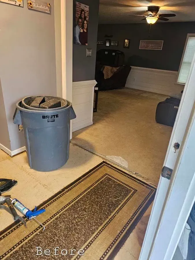 A room with a gray trash can, rug, and carpet before a renovation.
