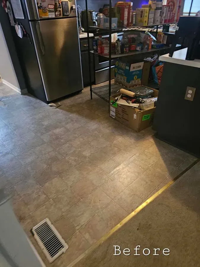 Kitchen with worn, light-colored flooring. Refrigerator, shelves with items, and cardboard boxes are visible.