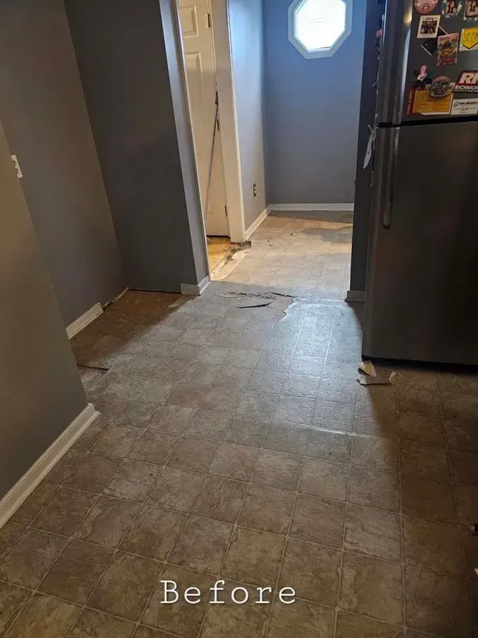 A worn tile floor in a kitchen with gray walls. A refrigerator and door frame are visible.
