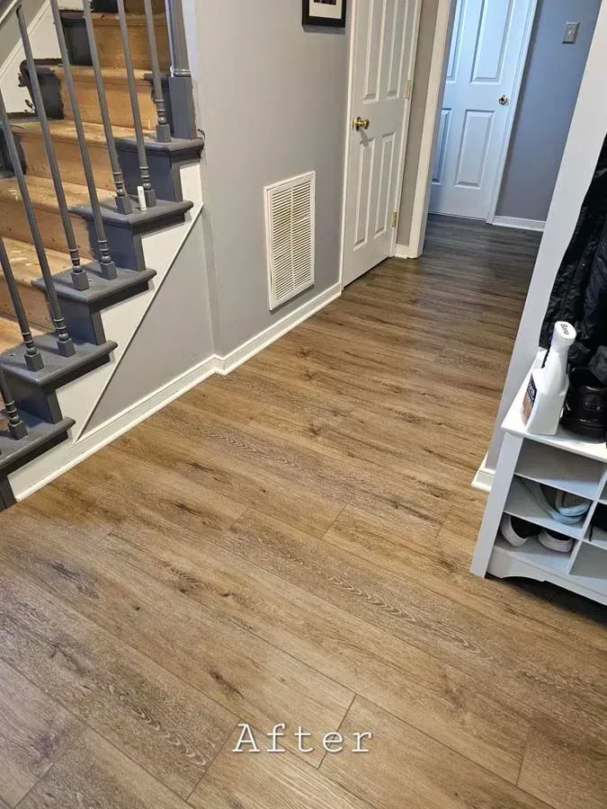 A renovated hallway with wood-look flooring and gray walls. A staircase, white trim, and shoe rack are visible.