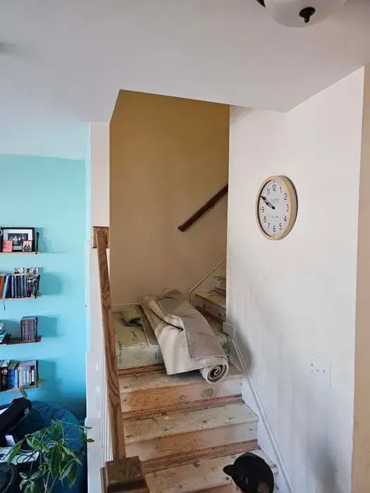 Staircase with carpet roll and mattress. Beige walls, wooden stairs, and a wall clock are visible.