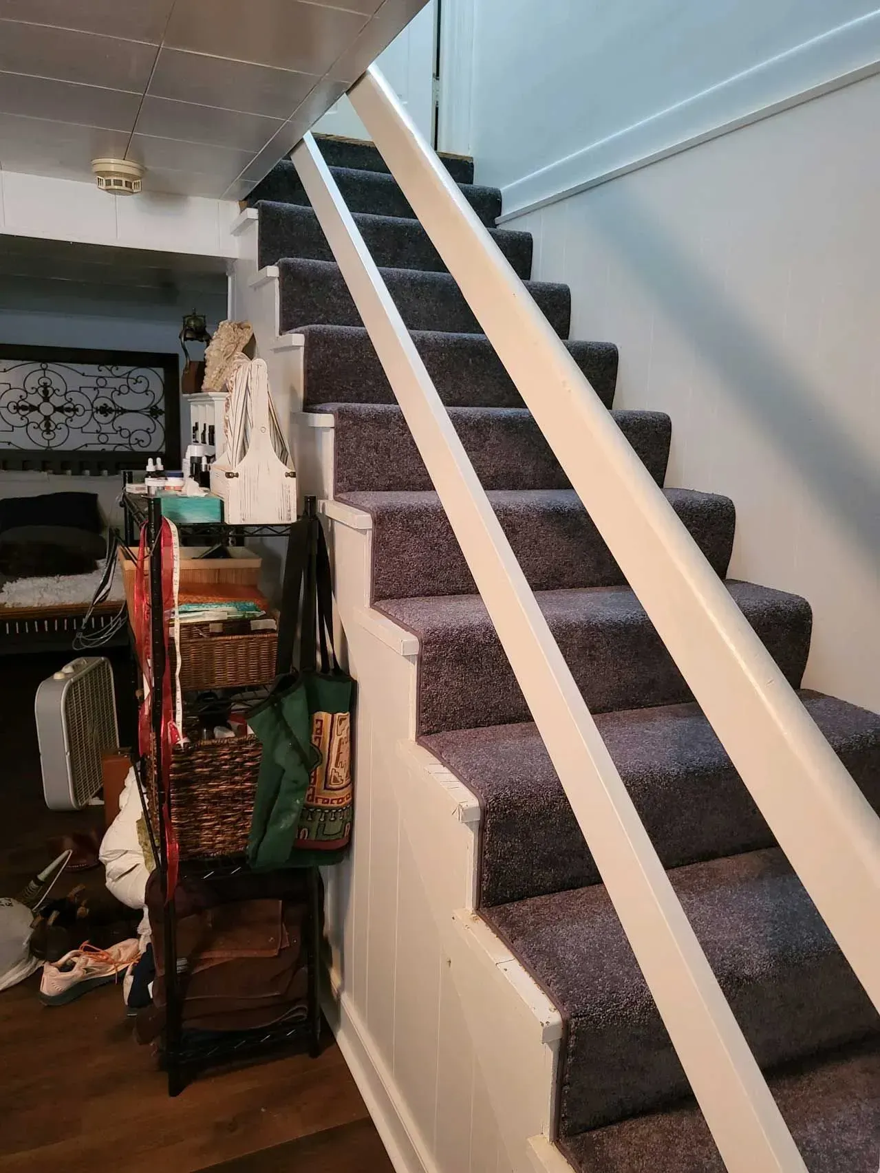 Staircase with gray carpet and white handrails. Items stored under the stairs: a fan, shelves, and bags.