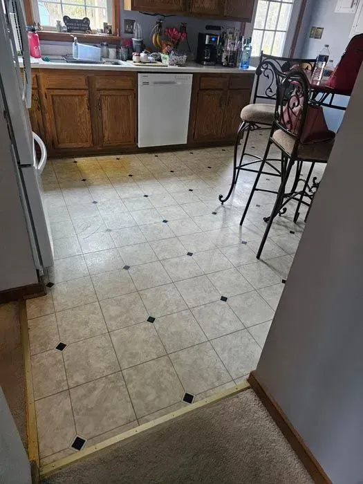 Kitchen with light-colored tile flooring, dark cabinets, and a white dishwasher. Two bar stools and a doorway visible.