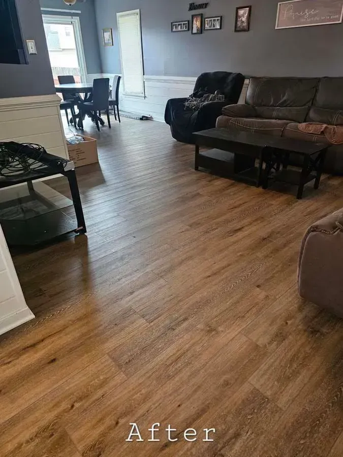 Living room with new wood-look flooring, furnished with black and brown furniture and a dining area visible.