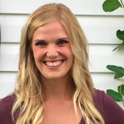 A woman with blonde hair is smiling and wearing a purple shirt.