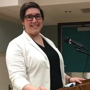 A woman in a white jacket is standing at a podium with a microphone.