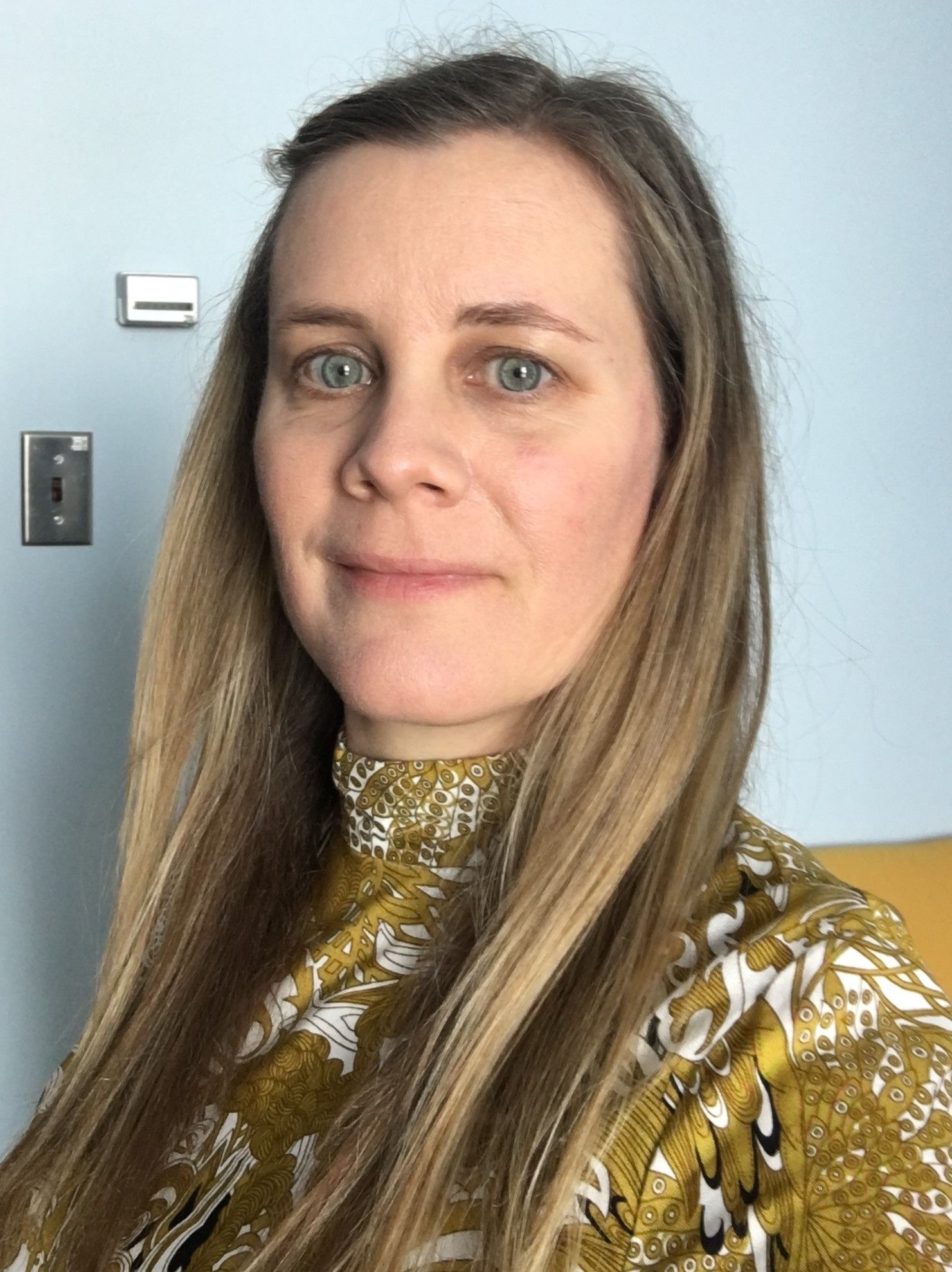 A woman with long blonde hair is wearing a yellow shirt and smiling for the camera.