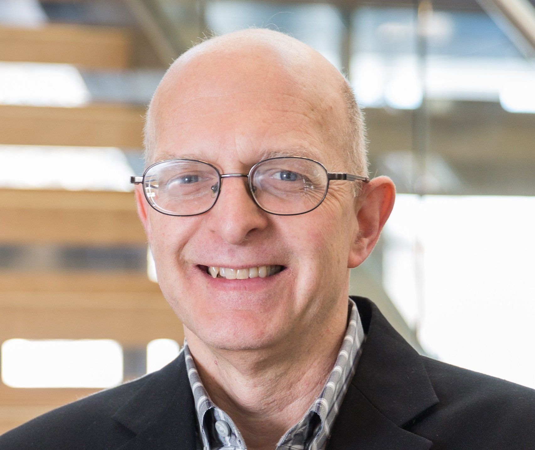 A man wearing glasses and a black suit jacket is smiling