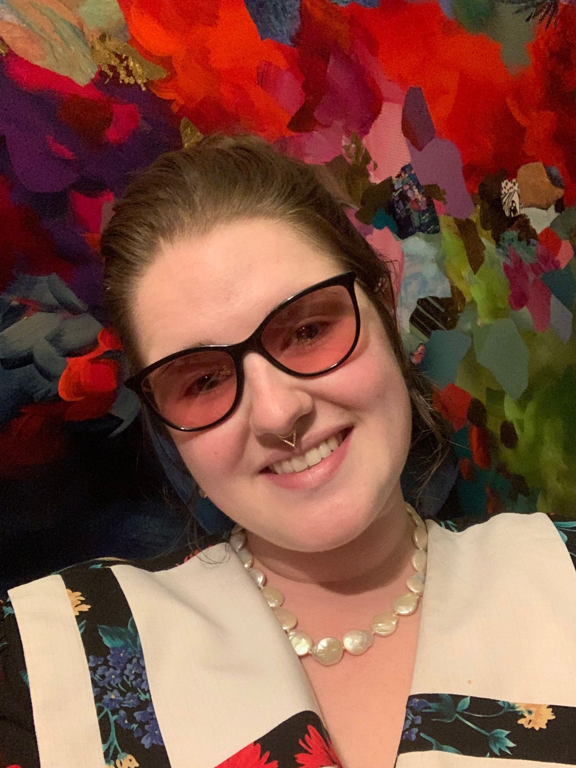 Woman standing in front of a colourful background wearing rose tinted glasses and smiling.