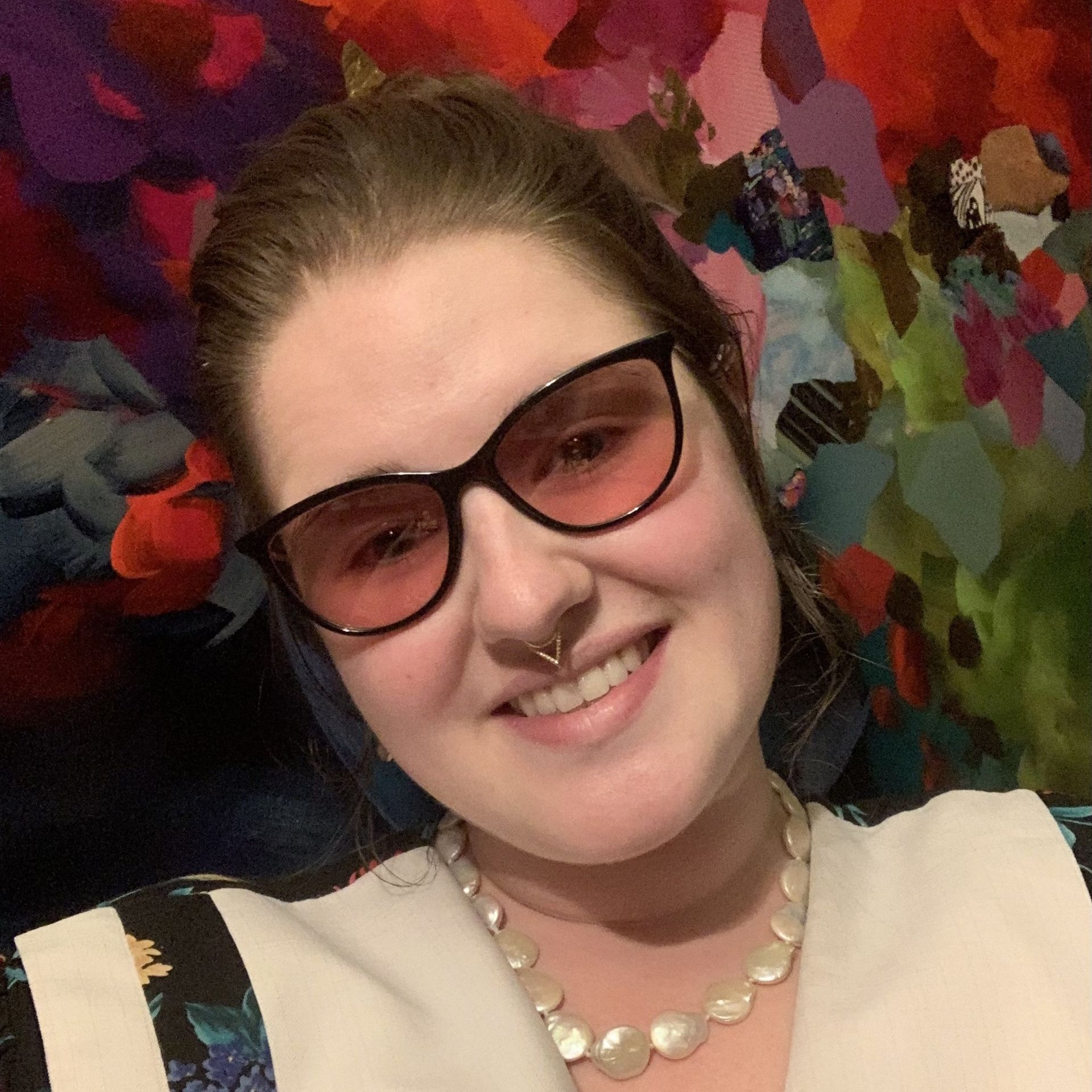 A woman wearing a necklace, nose ring and rose tinted glasses is smiling for the camera.