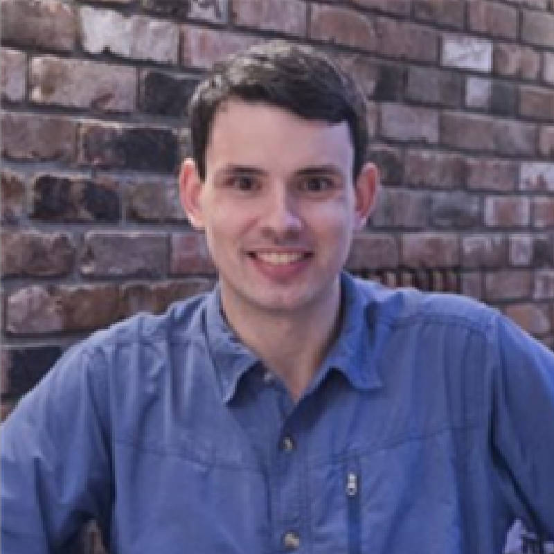 A man in a blue shirt is smiling in front of a brick wall