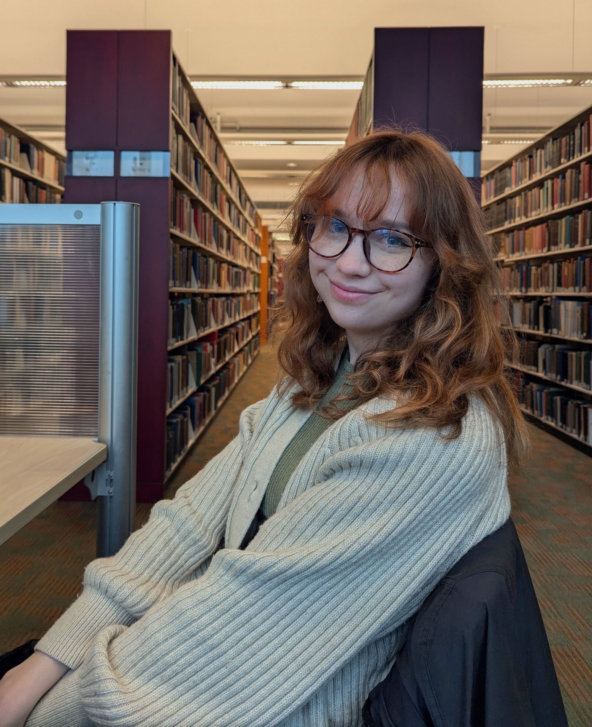 A portrait of a woman in a cream coloured cardigan sitting in a chair in a library and smiling at the camera.
