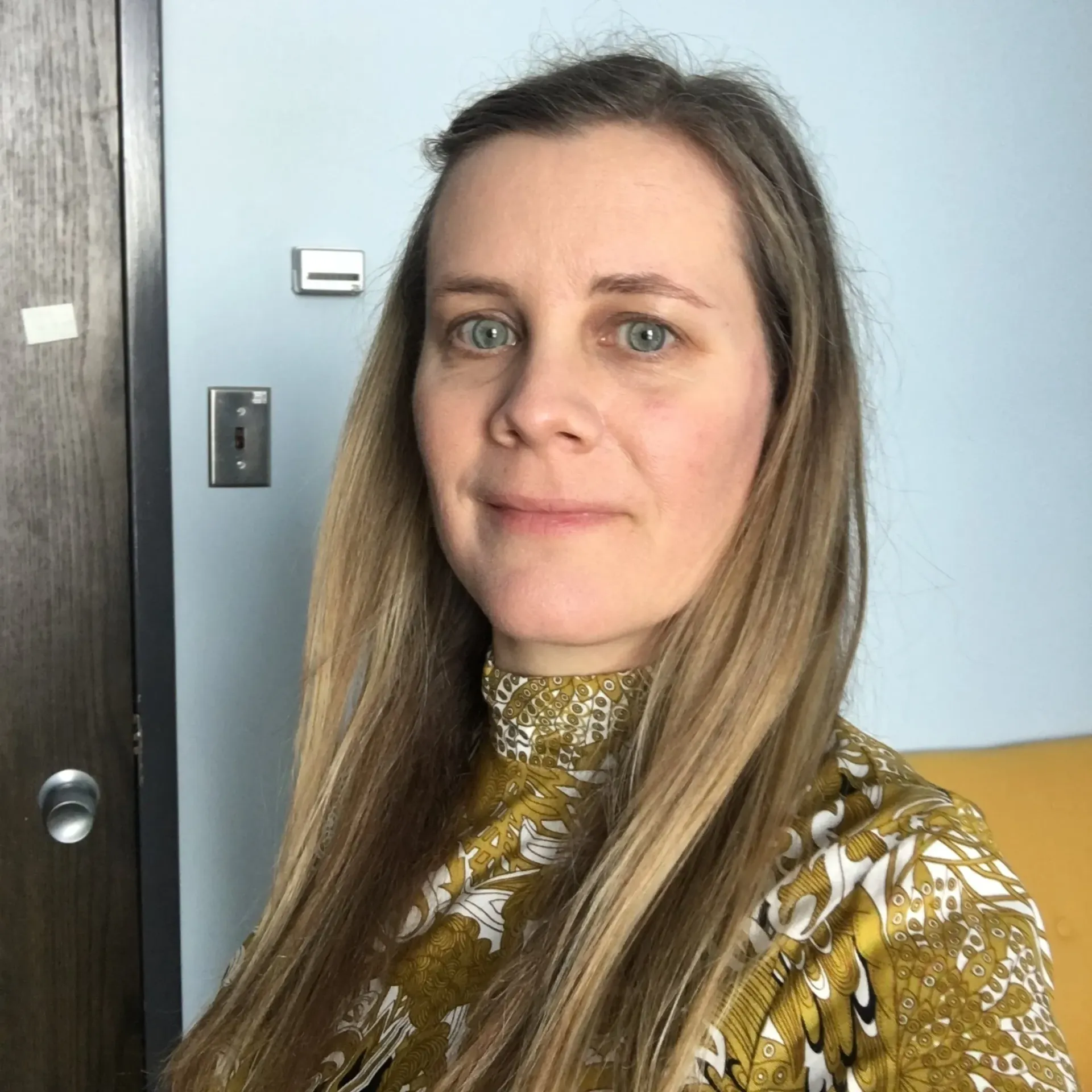A woman with long blonde hair is wearing a yellow shirt and smiling for the camera.