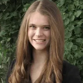 A young woman with long brown hair is smiling in front of a bush.