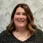 A woman in a black shirt is smiling in front of a tiled wall.