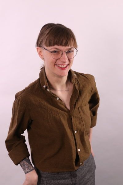 A woman with thin framed glasses and a brown button up shirt is smiling