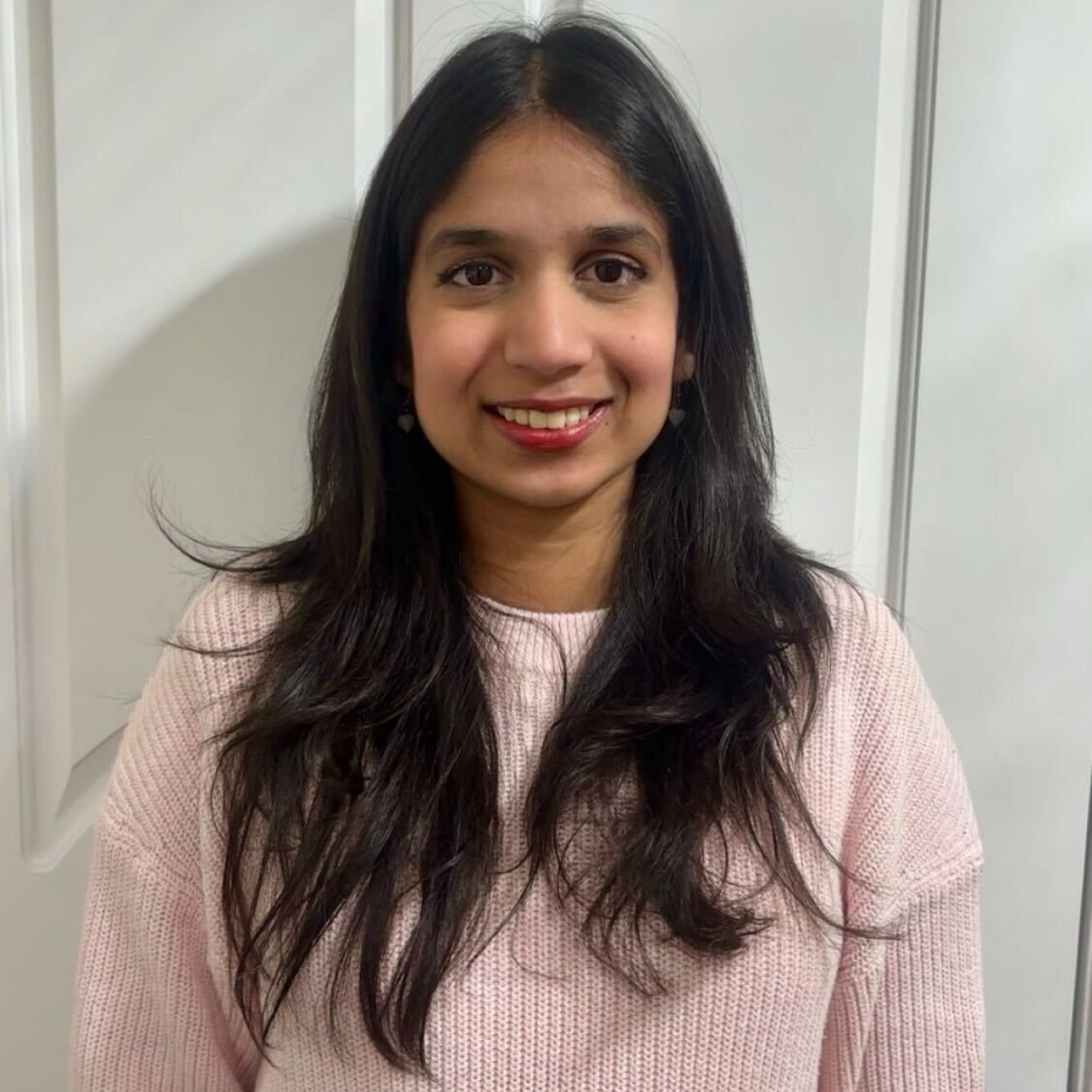A woman with long black hair and a pink sweater is smiling at the camera