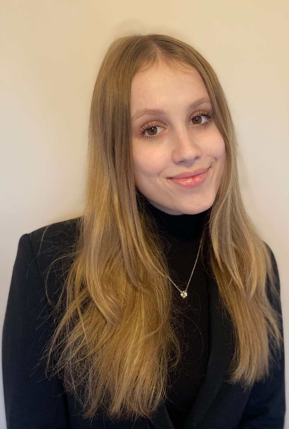 A woman wearing a necklace black tutrle neck and black blazer is sitting is looking at the camera and smiling.