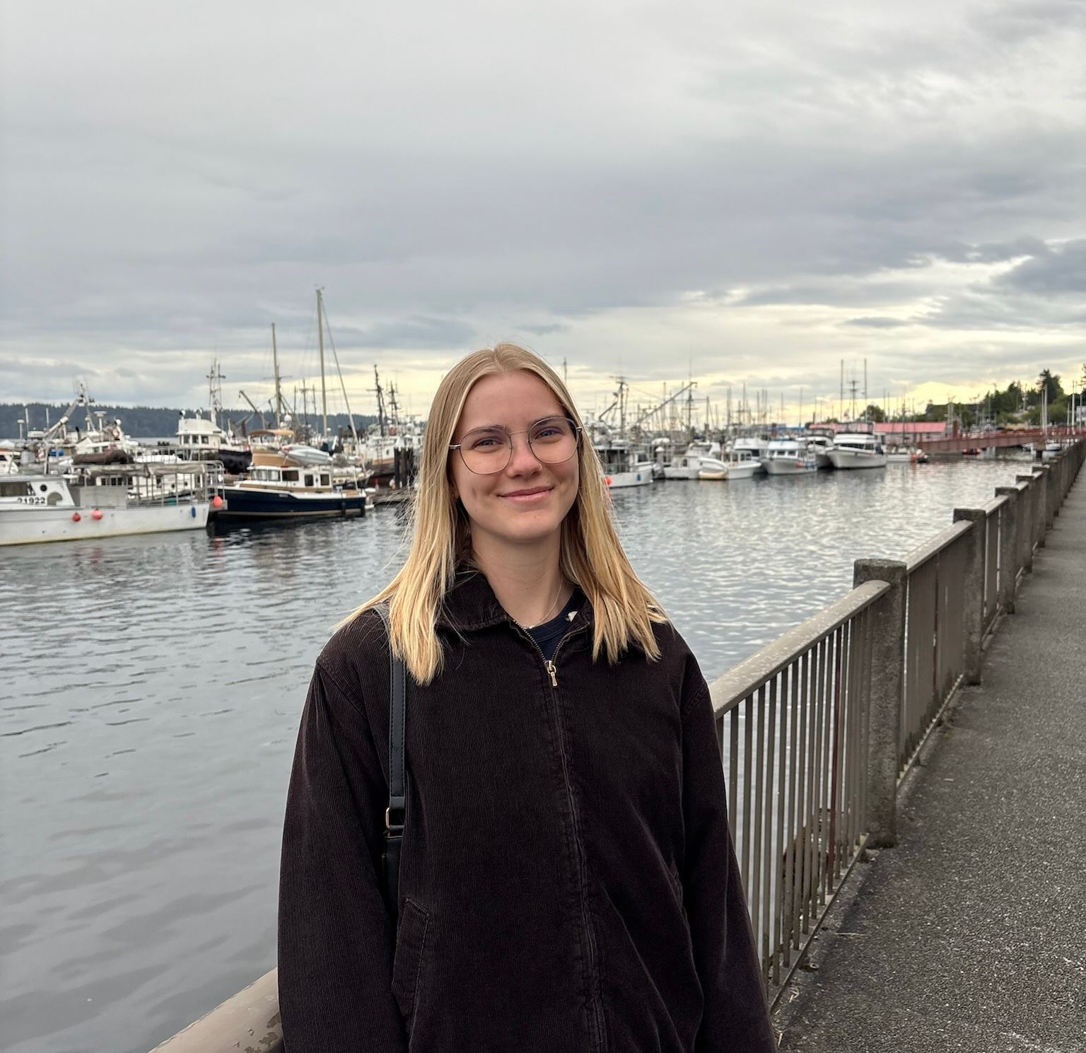 A woman in a black jacket is standing in front of a railing bordering a harbour.