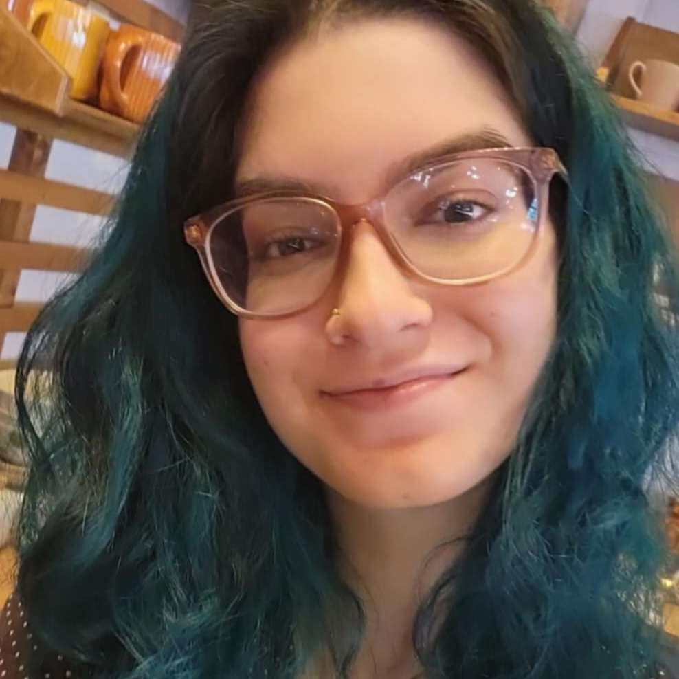 A woman with dyed teal hair and eyeglasses is smiling for the camera.