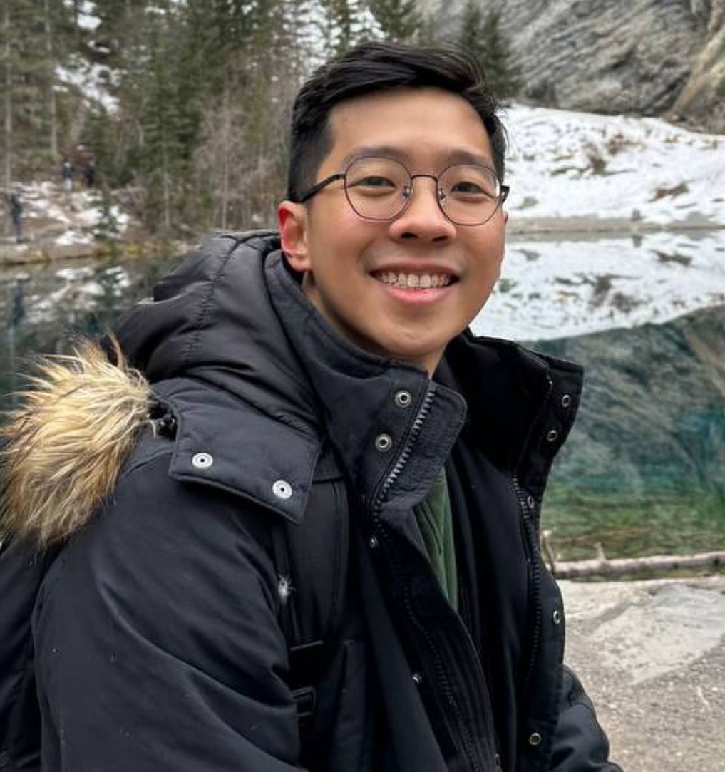 A young man wearing glasses and a black jacket with a fur hood is smiling.